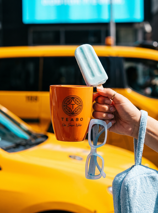 Une main tient un sachet Earl Grey Bio au-dessus d’une théière et d’une tasse en verre